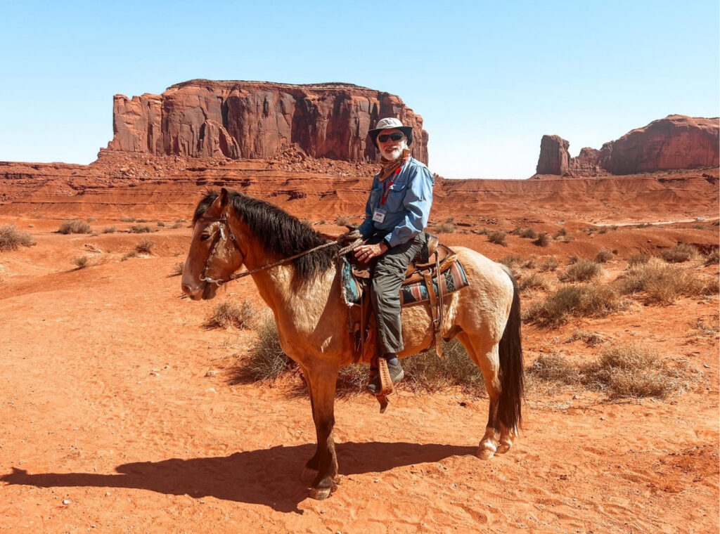 Rider on a blue-shirted person wearing a hat, sunglasses, and scarf, on a light brown horse in a red desert with sandstone cliffs behind.
