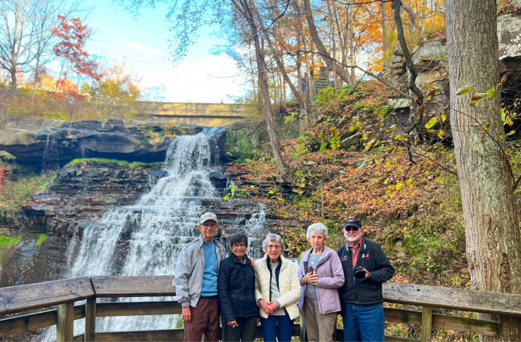 Five seniors stand together on a wooden viewing platform beside a multi‑tier waterfall with autumn trees in the background.