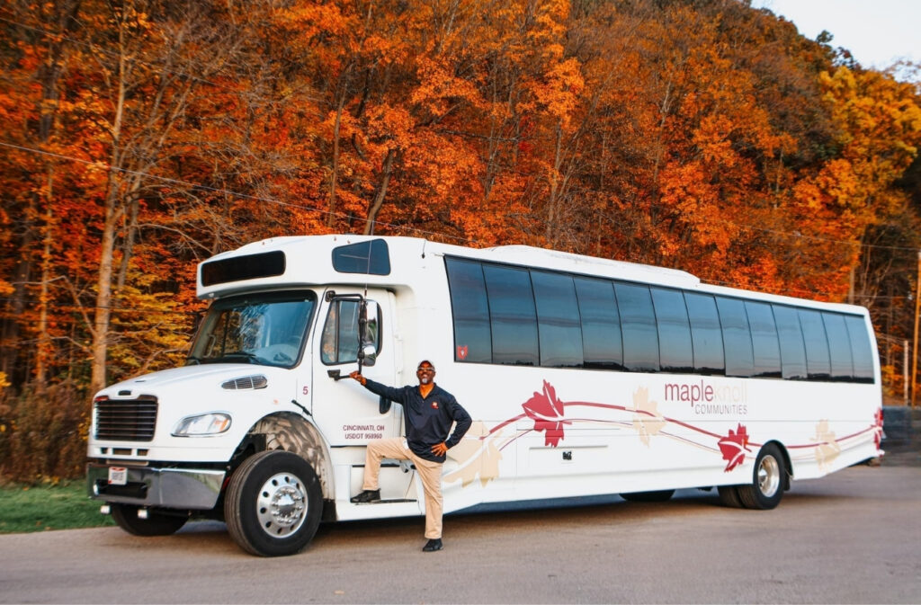 White tour bus with maple leaf graphics parked on a road, a man in front posing by the door, and orange autumn trees in the background.