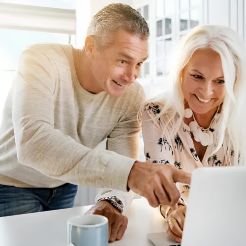 Smiling couple standing in a bright kitchen, pointing at a laptop on the counter.