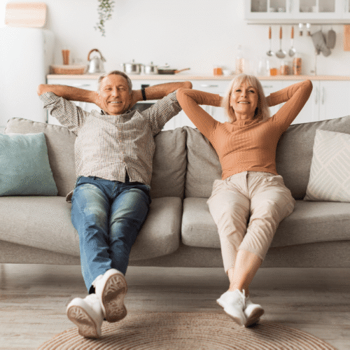 Older couple lounging on a gray sofa with arms behind heads, smiling in a bright, cozy living room with a plant nearby and a kitchen in the background.